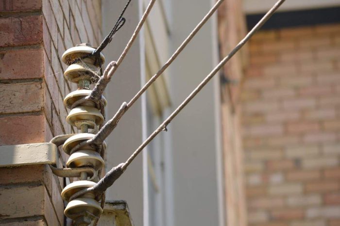 Ageing electricity wires An set of electricity wires are attached to a cobwebbed metal rod mounted on the side of a brick building.